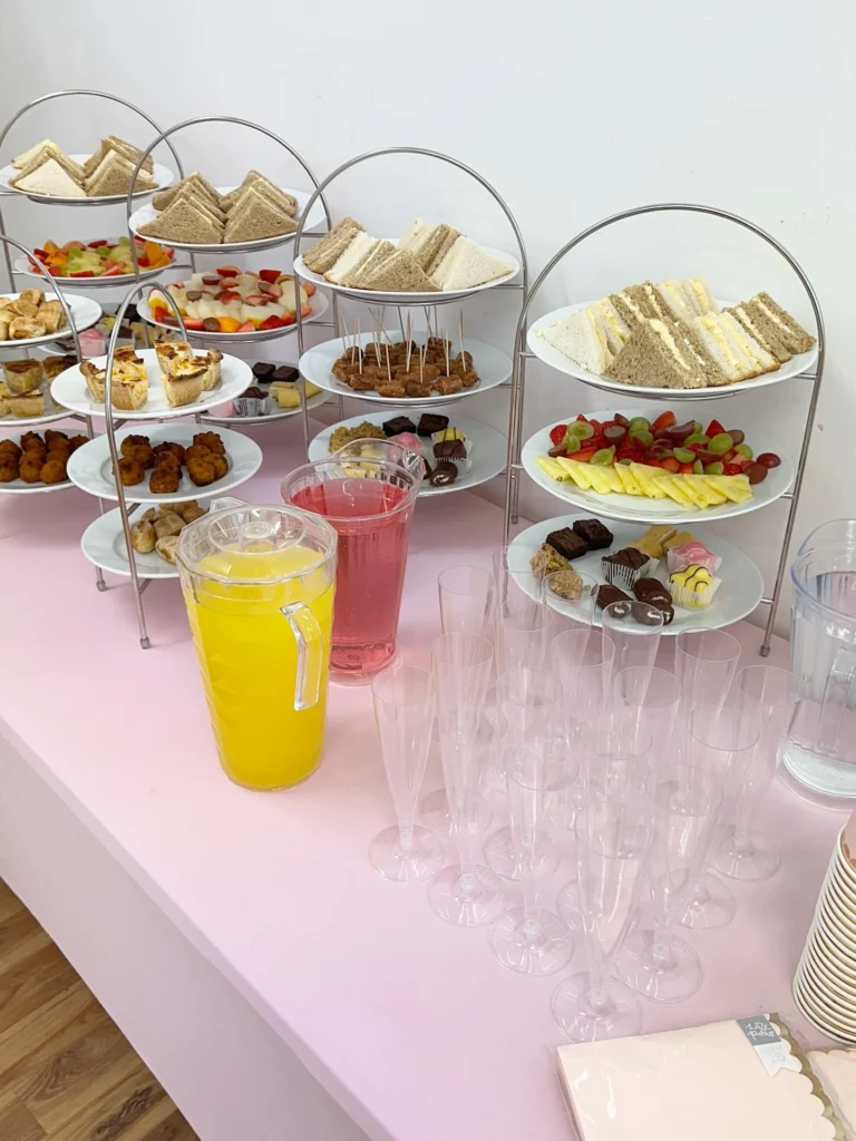 A buffet table with a pink tablecloth featuring tiered silver stands filled with finger sandwiches, fresh fruit, small pastries, and brownies, alongside pitchers of orange and pink juice and empty plastic champagne flutes.