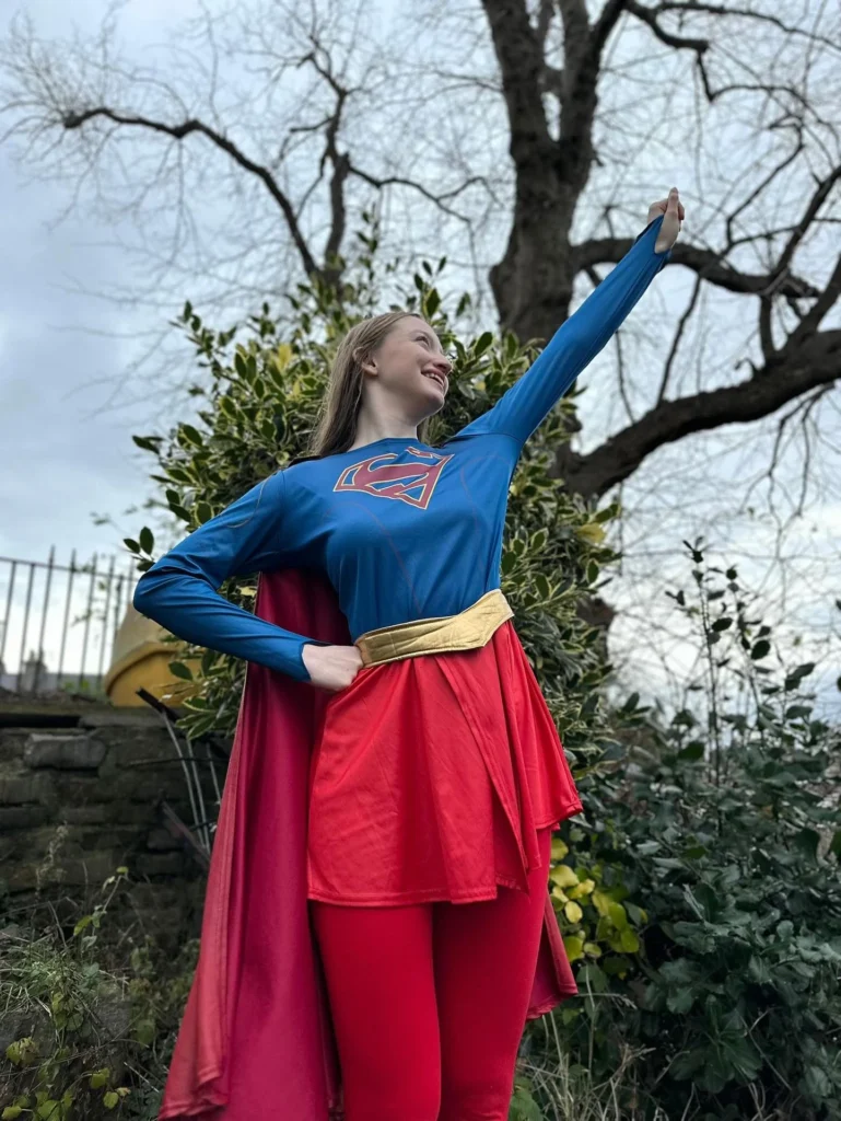 A young volunteer in a blue and red superhero suit with a cape, posing heroically with one arm raised in an outdoor garden setting. - Superheroes - Comics