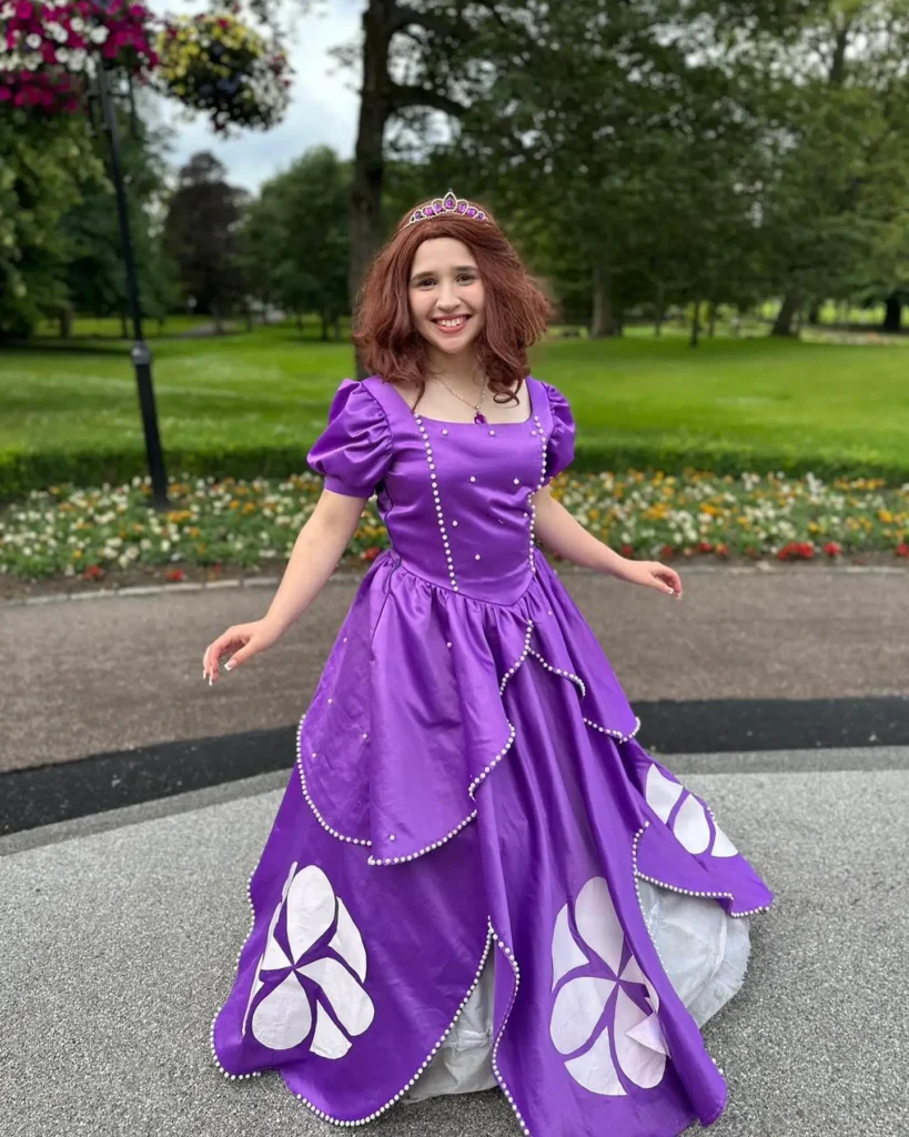 A smiling performer with auburn hair wearing a bright purple satin ballgown with white floral patterns and pearl detailing, wearing a small purple tiara and amulet necklace in a park.