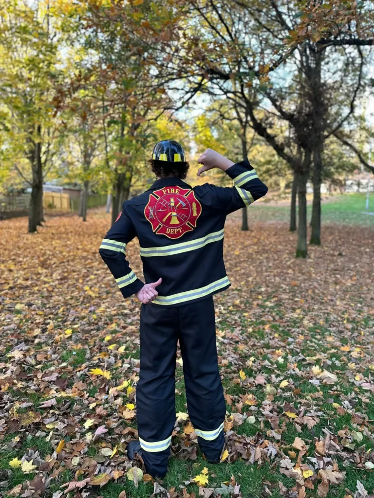 A performer from behind wearing a black fire fighter uniform with yellow reflective stripes and a "Fire Dept" emblem on the back. He is standing in an Aberdeen park filled with autumn leaves, wearing a black and yellow fire helmet.