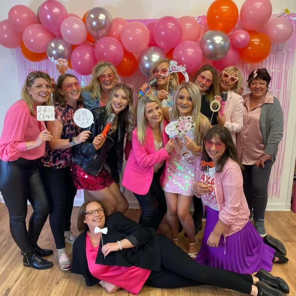 A group of cheerful women celebrating at an adult party, posing with pink and orange balloons, "Bride to Be" props, and microphones. They are dressed in vibrant pink and black outfits, with one woman reclining in the foreground holding a bow-tie prop.