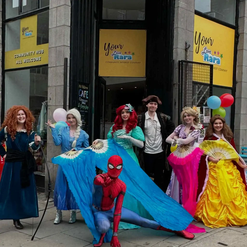 A group of seven performers in high-quality costumes standing outside the Your Love Rara Community Hub in Aberdeen. Characters include a Scottish princess with curly red hair, a snow queen, a mermaid with large blue fans, a superhero in a red and blue suit, a pirate, and two princesses in purple and yellow ballgowns.