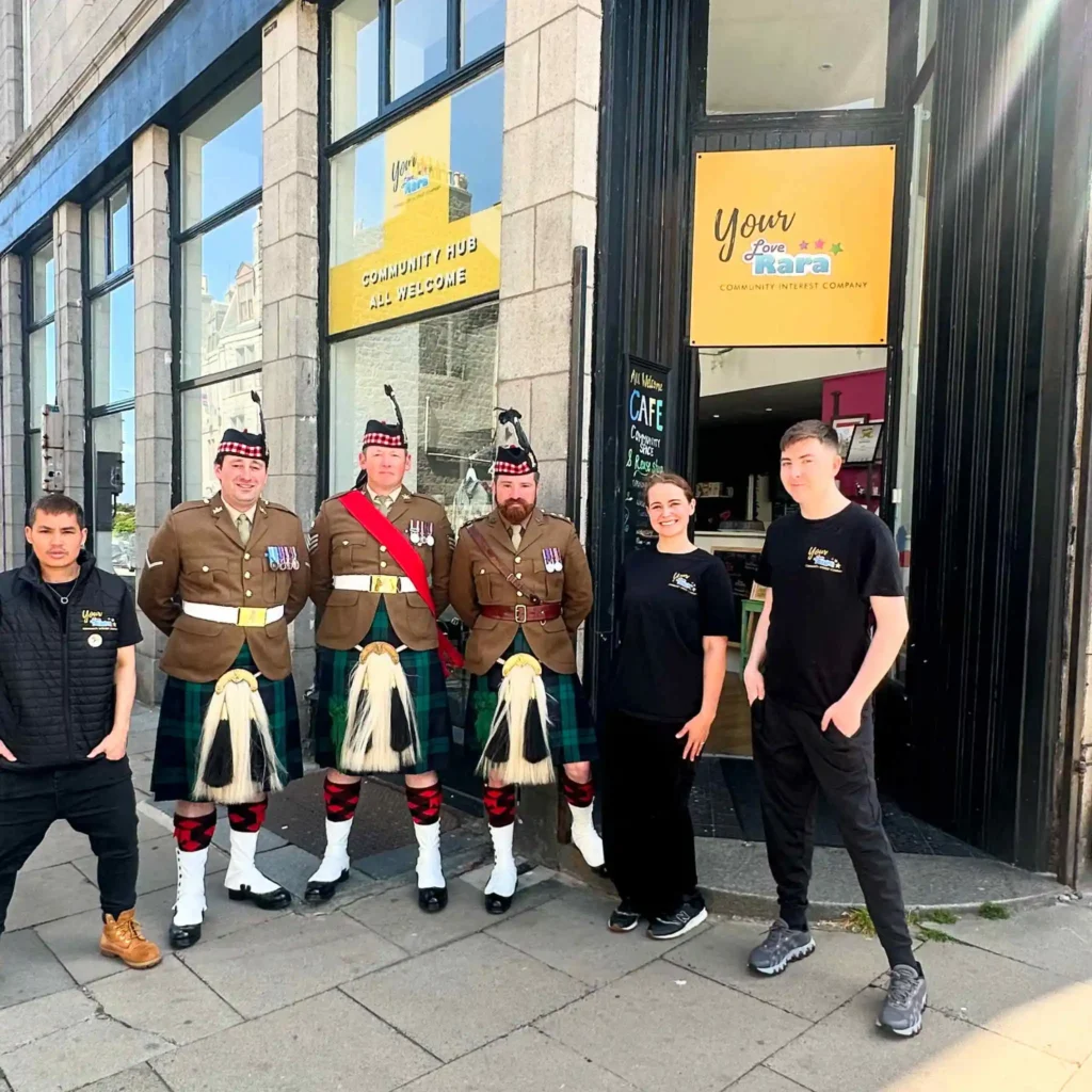 Three men in traditional Scottish military Highland dress, including kilts and sporrans, standing outside the Your Love Rara Community Hub in Aberdeen alongside three staff members in branded black t-shirts.