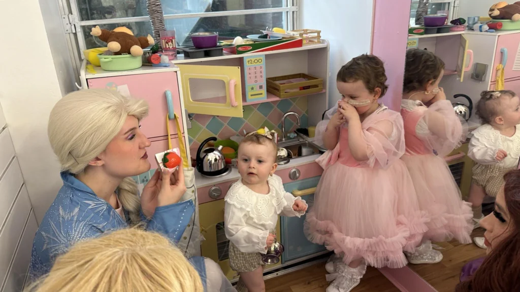 A performer dressed as a snow queen in a blue dress interacts gently with a toddler in a white blouse. To the right, a young girl in a pink princess dress stands by a toy kitchen set, looking into a mirror. The setting is a bright, colourful play area at the Your Love Rara Hub.