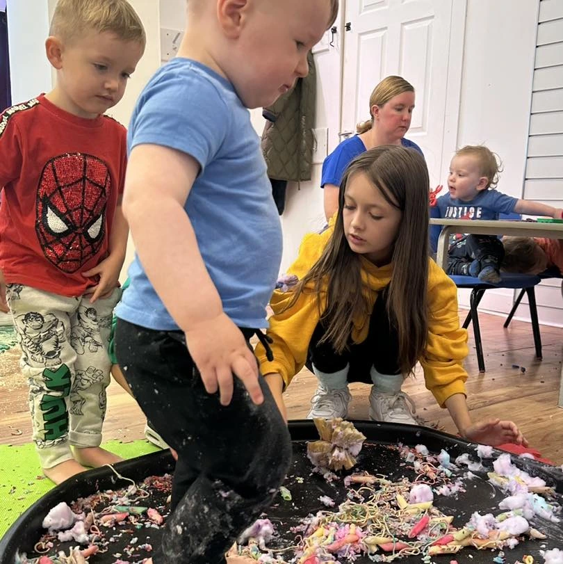 Children and a young volunteer in a yellow Your Love Rara hoodie engaging in "messy play" around a black sensory tray filled with colourful materials like pasta and foam. A young boy in a blue t-shirt stands in the tray exploring the textures.
