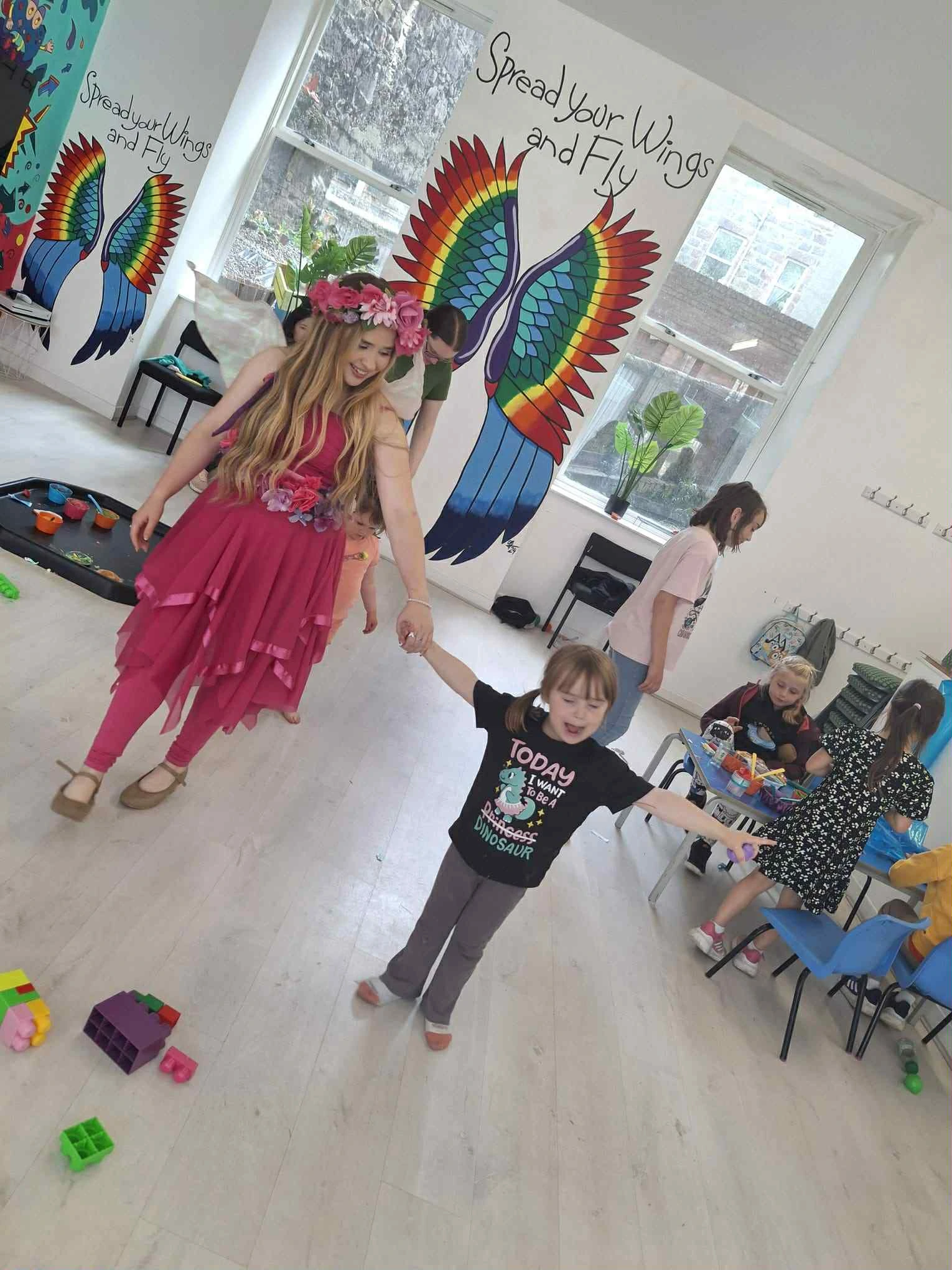 A performer in a pink fairy costume with a floral crown holding hands with a young girl in a "Princess Dinosaur" t-shirt at the Your Love Rara Hub. In the background, other children are engaged in messy play and crafts near large colourful wing murals.