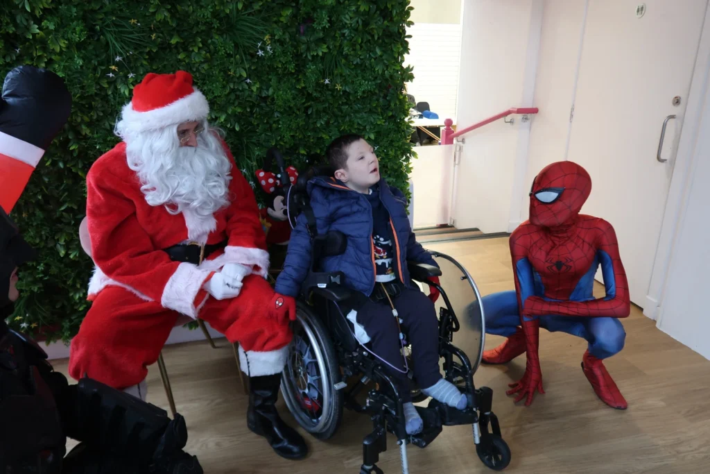 A young boy in a wheelchair sitting between Santa Claus and a performer dressed as Spider-Man. They are in a room with a green leafy wall backdrop, creating a joyful and inclusive holiday moment.