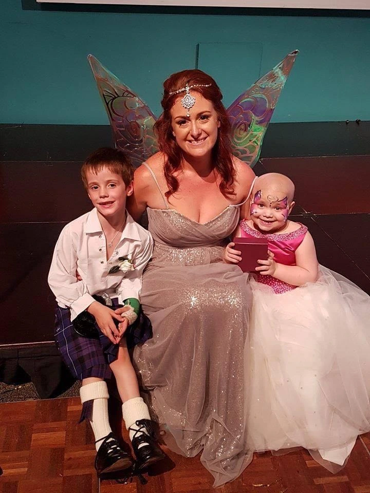 A smiling female performer (Zara Grant) dressed as a fairy with large iridescent wings and a jewelled headpiece. She is seated between two children: a young boy in a traditional Scottish kilt and a young girl in a pink and white dress with butterfly face paint.