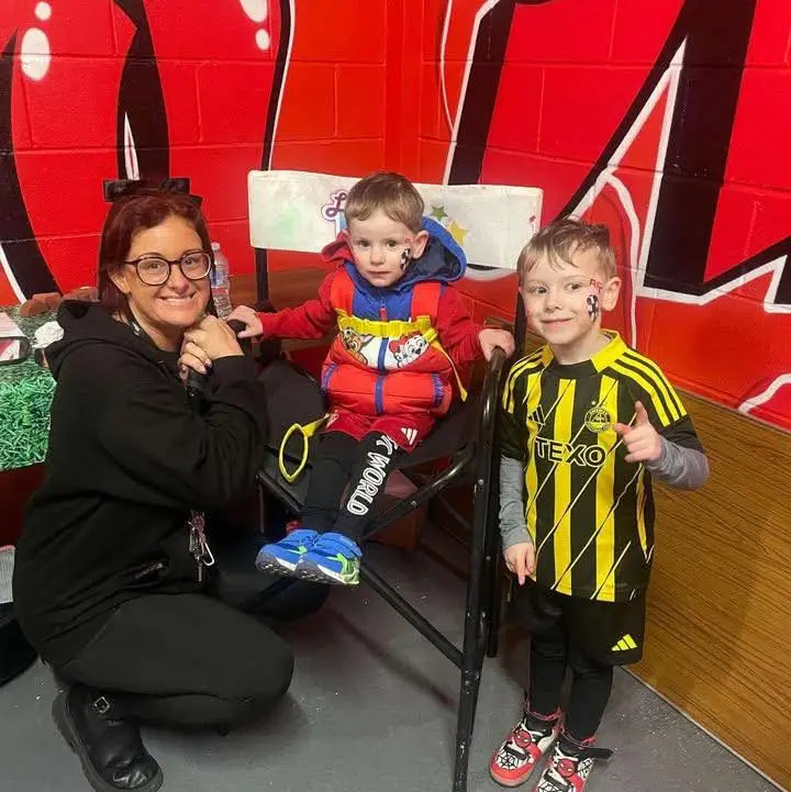 A staff member in a black Your Love Rara hoodie kneeling next to two young boys. One boy is seated in a director's chair wearing a red gilet, and the other is standing in a yellow and black Aberdeen FC football shirt. Both boys have decorative face painting designs on their cheeks.
