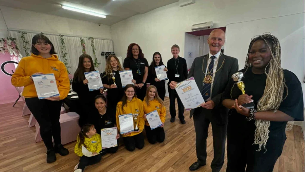 A group of young people and adults at the Your Love Rara Hub, holding Saltire Award certificates and silver trophies. They are standing alongside a man in a formal suit and mayoral chain of office. Several young people are wearing yellow Your Love Rara hoodies.