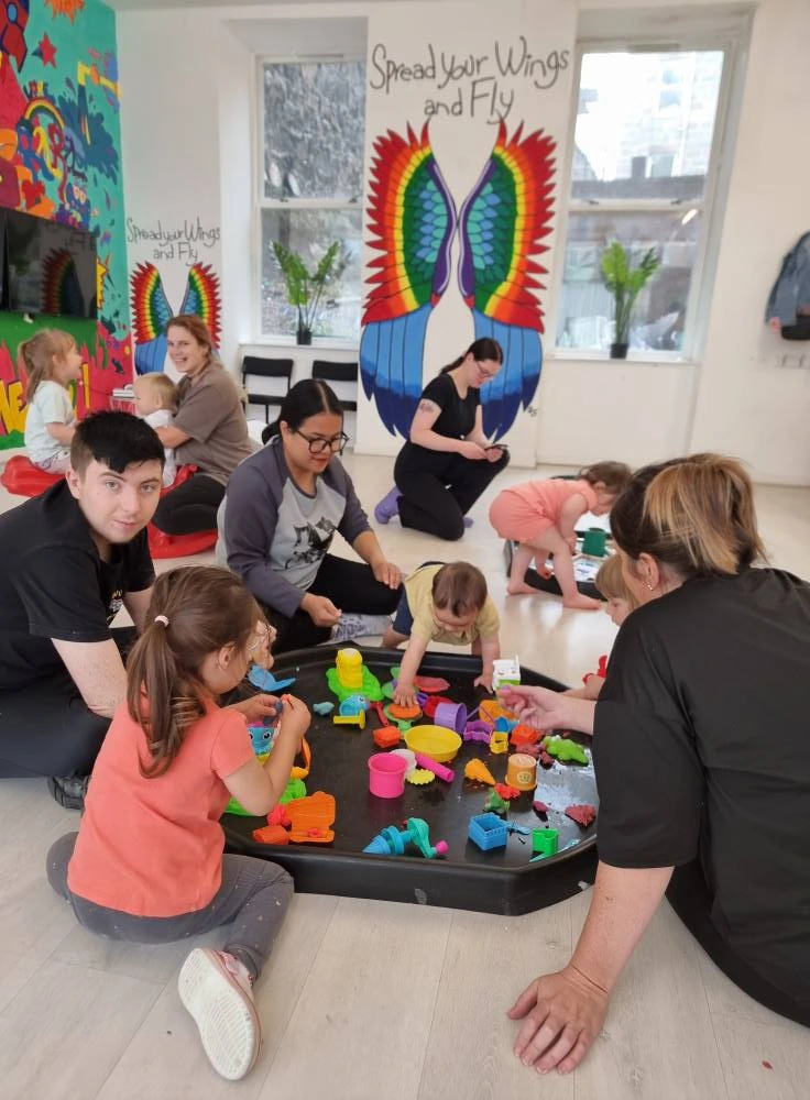 A group of children and adults sitting on the floor around a large black sensory tray filled with colourful toys. In the background, there is a white wall with a large mural of colourful wings and the words "Spread Your Wings and Fly," and another vibrant, multi-coloured mural.