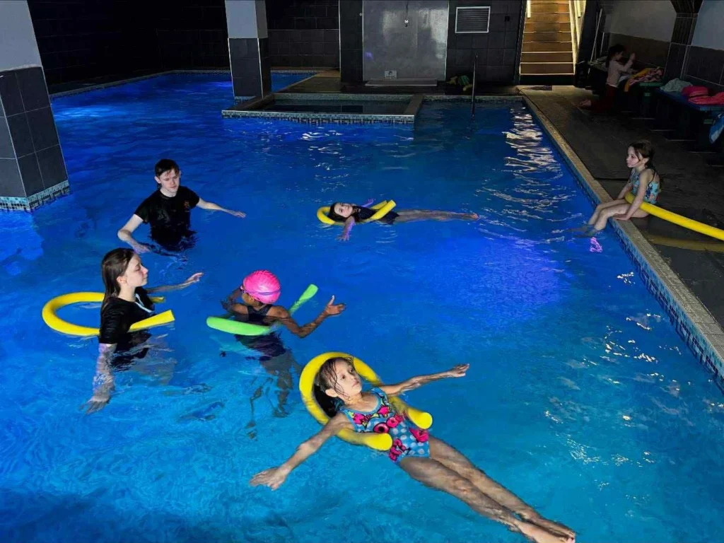 A group of children enjoying a swimming session in a blue-lit indoor pool. Some children are floating using yellow and green foam noodles, while others are interacting with a staff member in the water. One child sits on the edge of the pool.