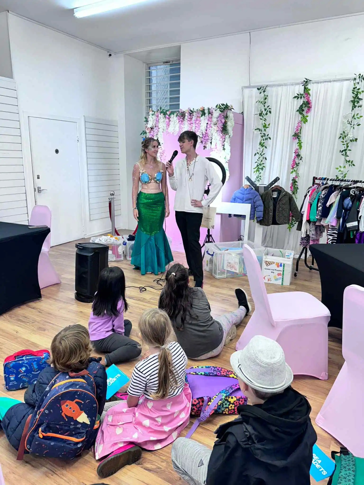 A performer in a green and blue mermaid costume with a seashell top stands next to a male performer in a white shirt and black trousers holding a microphone. They are performing for a group of children sitting on the floor at the Your Love Rara Hub, which is decorated with pink floral arrangements.