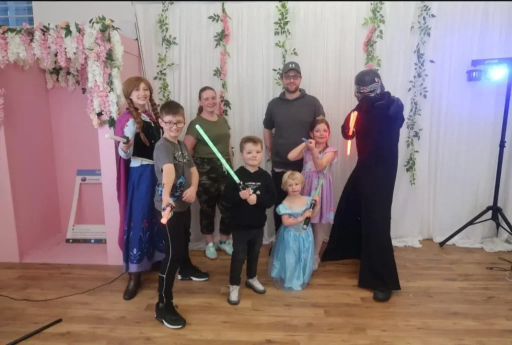 A group of children and adults posing with characters in a decorated studio at the Your Love Rara Hub. Characters include a princess in a blue and purple dress and a dark-clothed sci-fi villain with a red lightsaber. The children are holding glowing toy sabers against a backdrop of white curtains and floral arrangements.