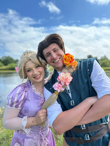 A smiling female performer dressed as a blonde princess in a lavender lace dress and crown, standing next to a male performer dressed as a roguish thief in a teal buckled vest. The princess is holding her long blonde braid, which is decorated with pink and orange flowers and draped over the man's shoulder. They are outdoors with a river and blue sky in the background.